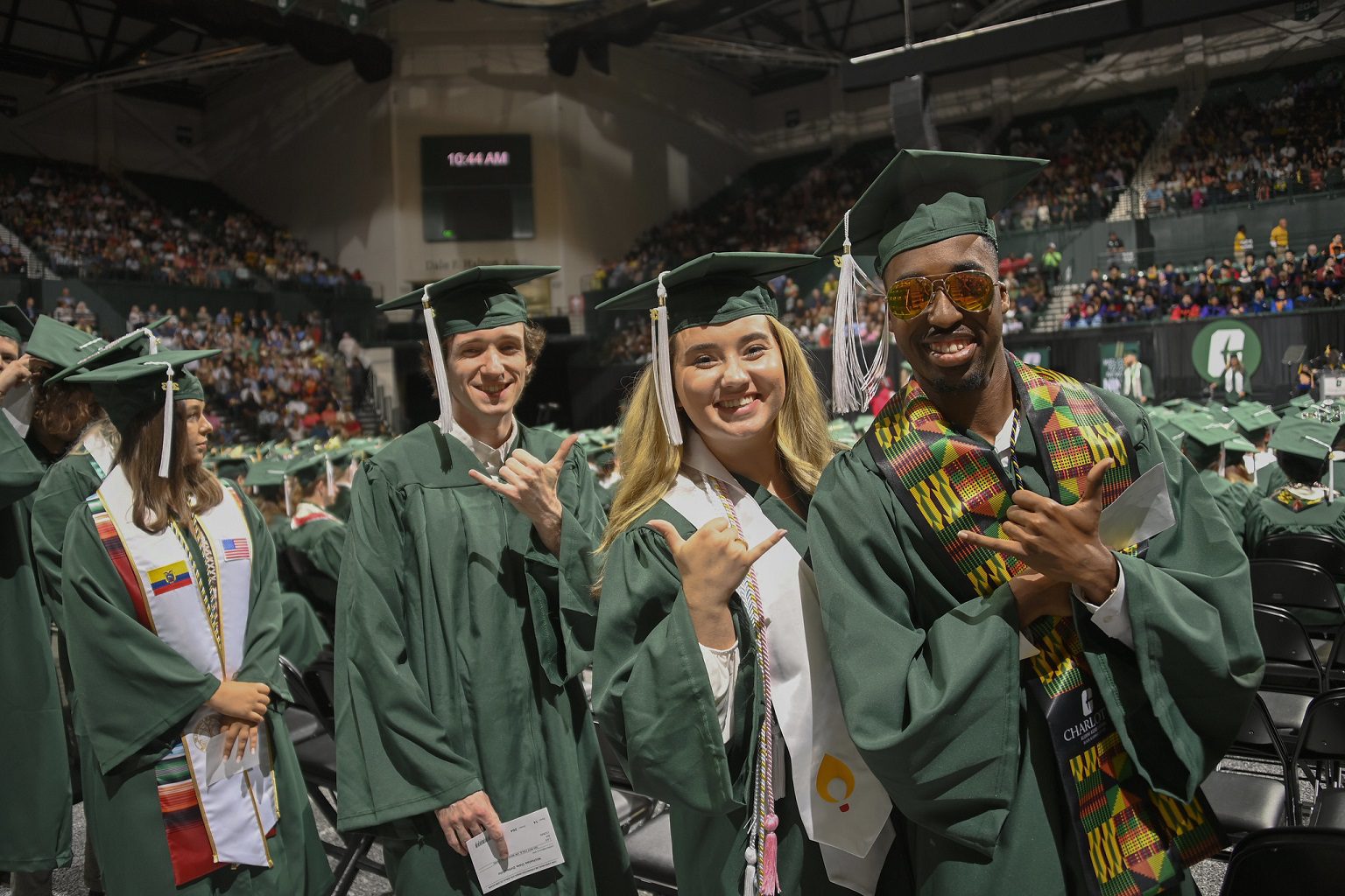 Three graduates in caps and gowns smile into the camera while holding their hands in the Charlotte pick-axe symbol. 