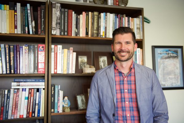 Jason Black with books on shelves behind him. Dark haired man with a beard and mustache wearing a red plaid shirt and blue coat.