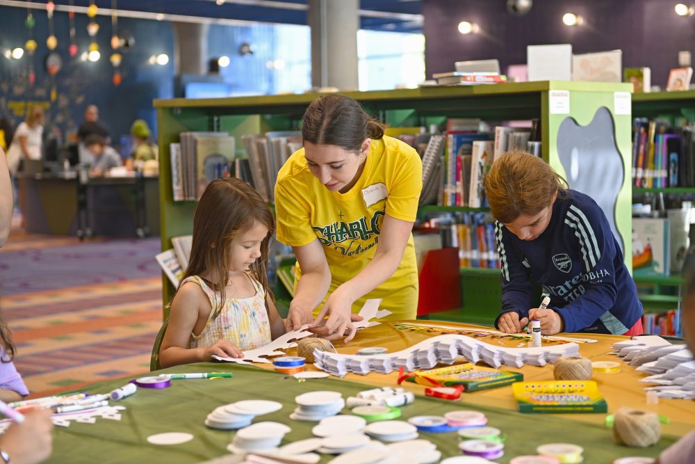 A UNC Charlotte student volunteer in a bright yellow CharlOz shirt helps kids make crowns at the craft table.