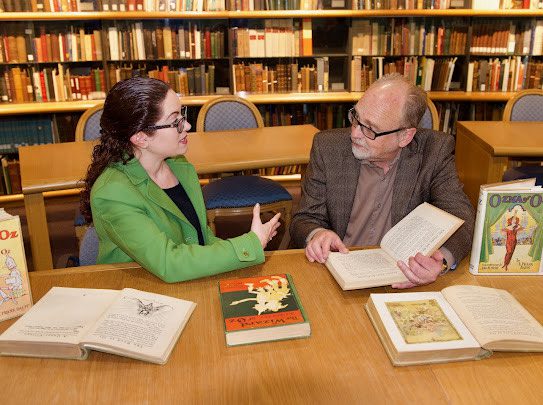 Dina Massachi and Mark West sit a table talking, surrounded by books about the Wizard of Oz.