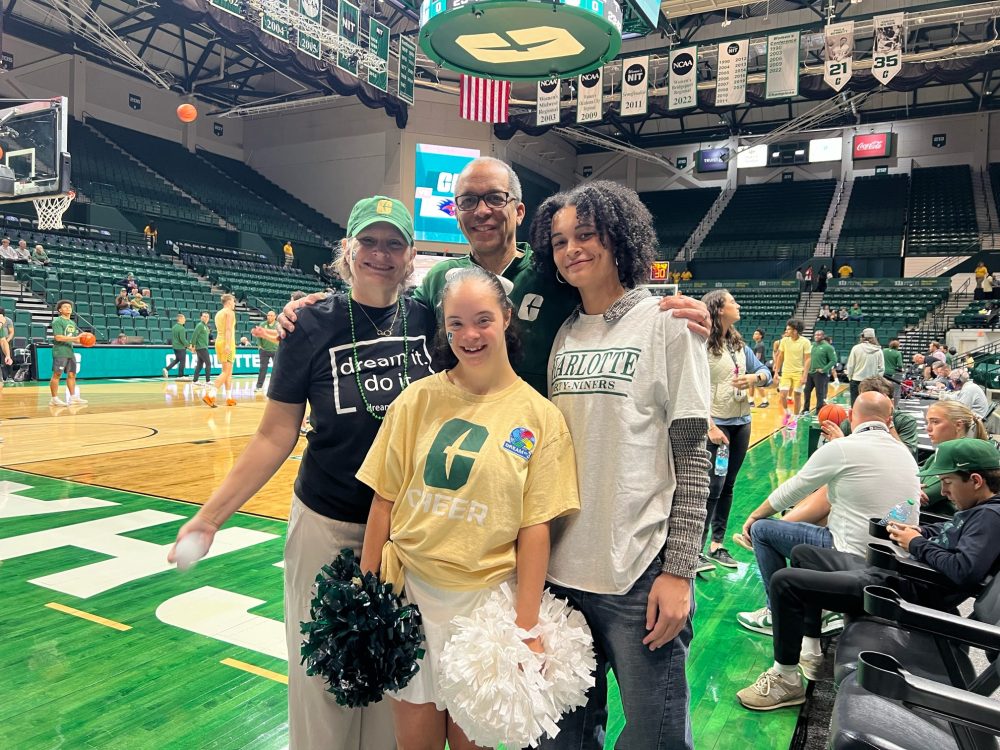 Maya Barbee and her mom, dad and sister wear Charlotte gear and stand on the court at a Charlotte basketball game.