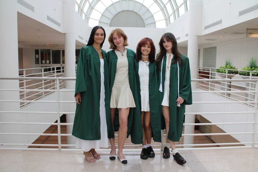 Four women wear green graduation gowns over white dresses, standing against a white railing in the atrium.