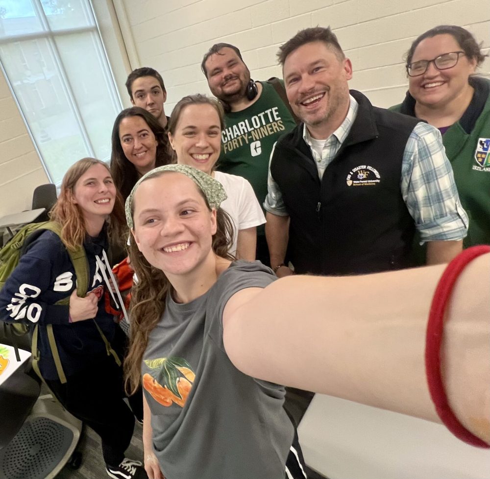 Hawkins stands smiling with a group of 7 fellow Niner Classmates, selfie style, with the student in front taking the photo.