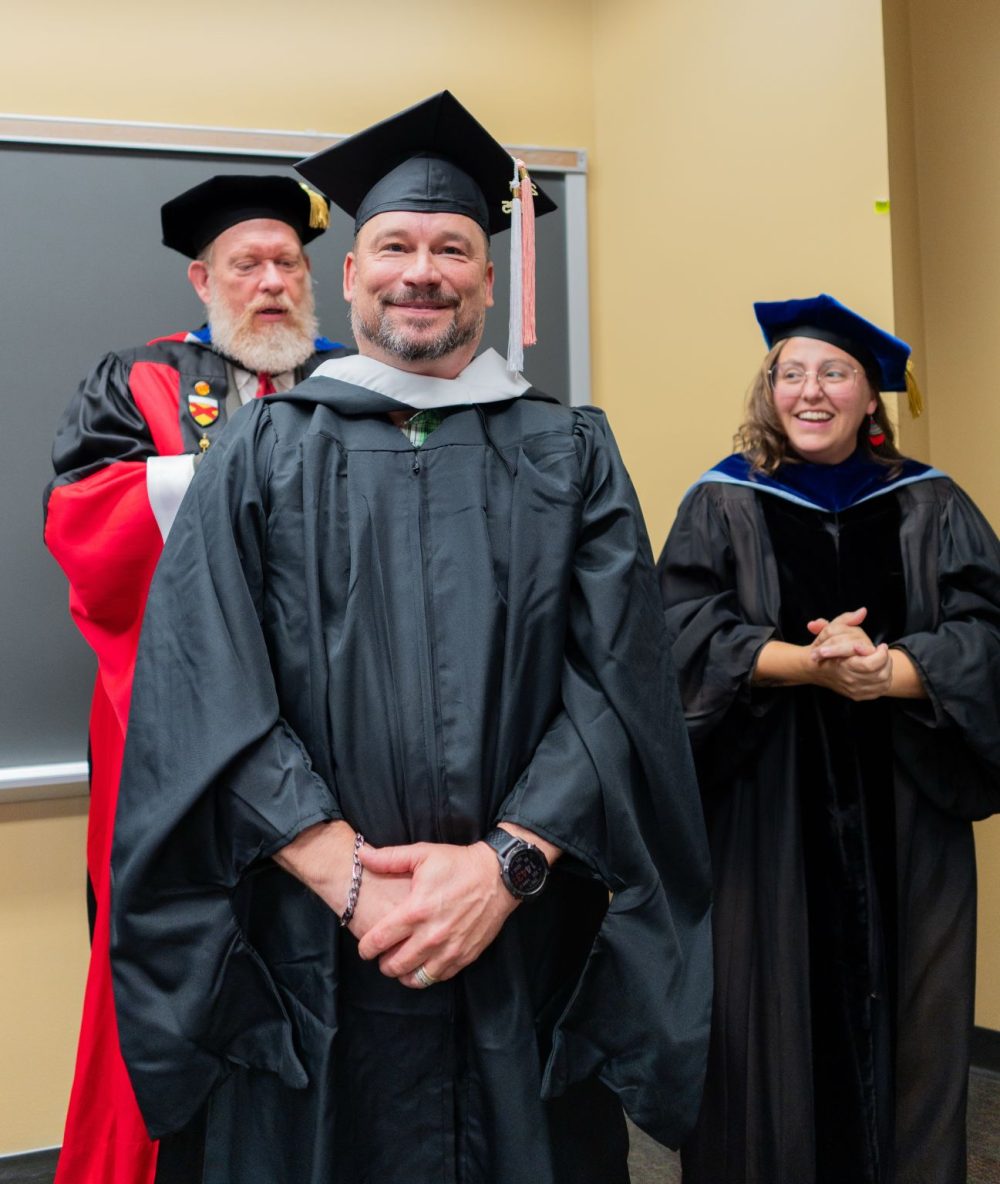 Hawkins smiles as he is hooded with two faculty members in regalia from his department.