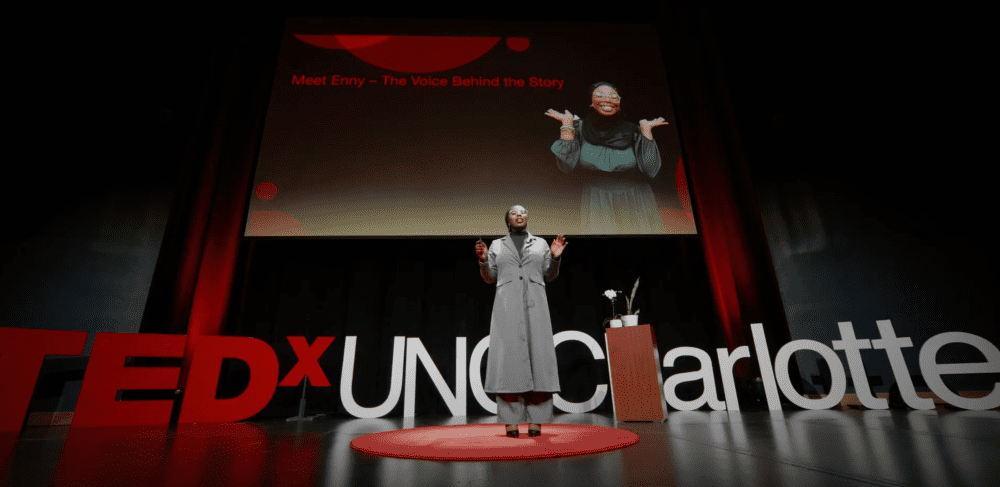 Enny on stage in front of the TEDx UNC Charlotte logo and her smiling on the screen behind her.