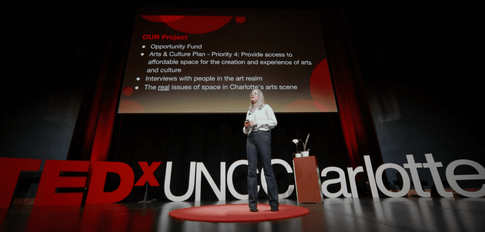 Jenna Drew on the TEDx UNC Charlotte stage with the logo and screen behind her.