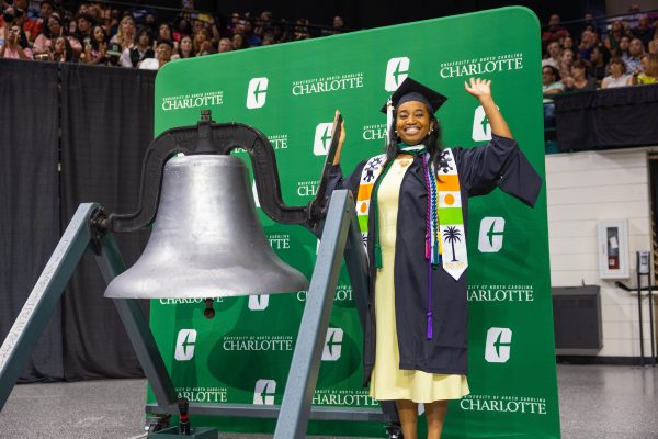 Pennda Arami wears graduation regalia, and smiles and waves while holding the handle of the large silver bell at UNC Charlotte's first-ever summer graduation.