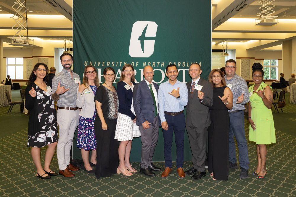 Ten of the newly promoted faculty members pose in front of the UNC Charlotte logo with Dean Chris Boyer in the middle. Most are doing the UNC Charlotte pickaxe sign with their hands.