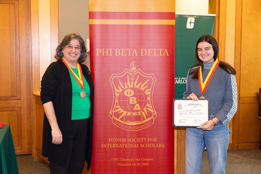 Hill posing at the Phi Beta Delta Mu induction ceremony alongside Adriana Medina and a PBD branded banner.