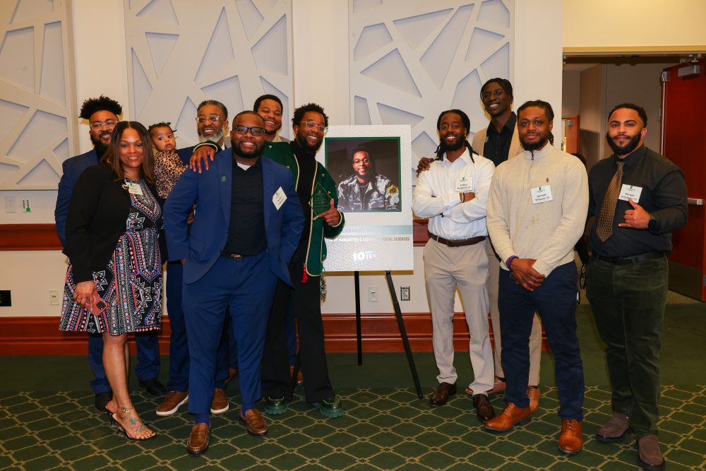 C. Emmanuel Wright poses with a large group of his supporters, standing around his award poster with his photo on it. 