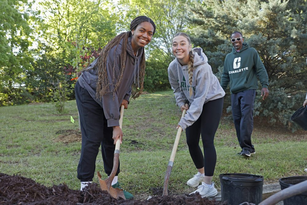 Two students smile at the camera while shoveling soil during a tree planting event for Spring Campus Beautification Day 2025. Another person smiles in the background.