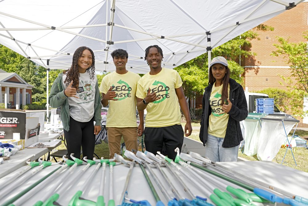 Four students smile at the camera while under a tent during Spring Campus Beautification Day 2025. Trash grabbers are placed on the table in front of them, and they are wearing t-shirts that say "Zero Waste"