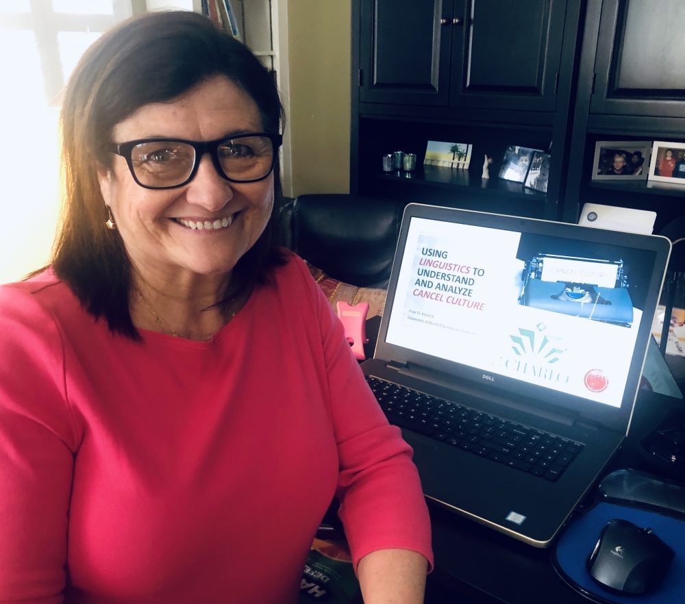 Blitvich posing in front of her laptop in her home office. A PowerPoint presentation is on the screen, title "Using linguistics to understand and analyze cancel culture"
