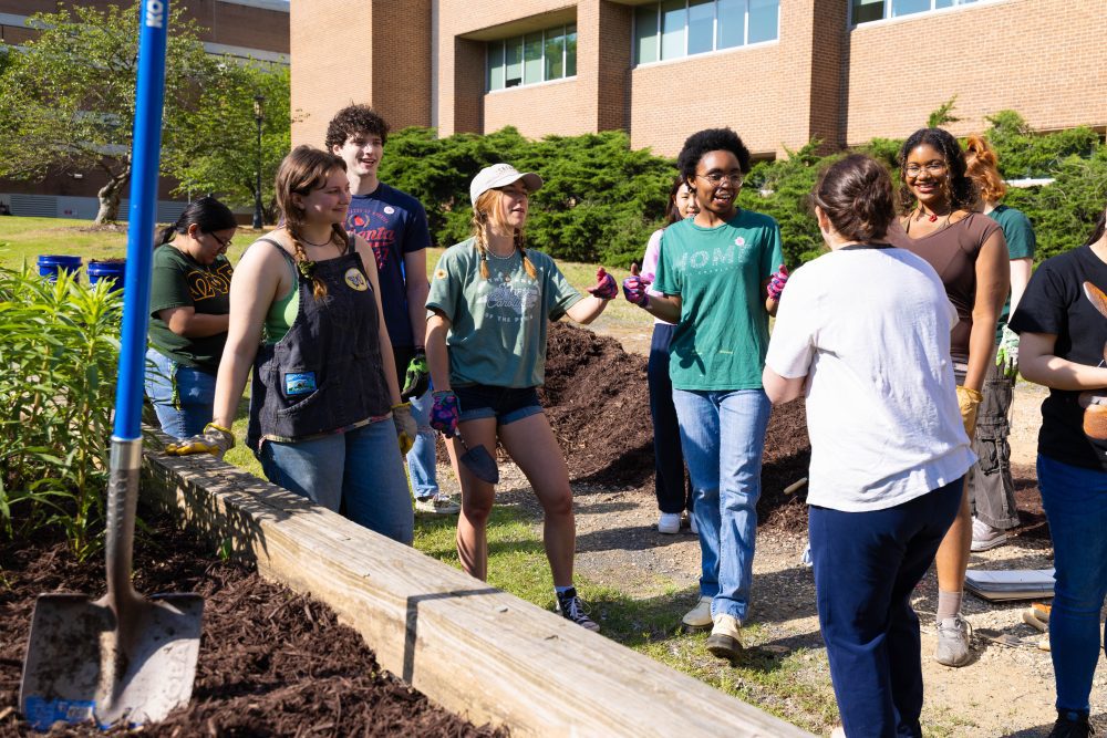 A group of students gather on Garden Day 2025, listening to an instructor. A shovel is placed in the garden bed in the foreground with a building in the background.