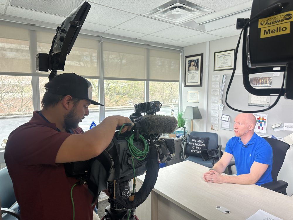 Horizontal image of Terry Shirley facing a cameraman operating a camera while sitting at his desk in his office.