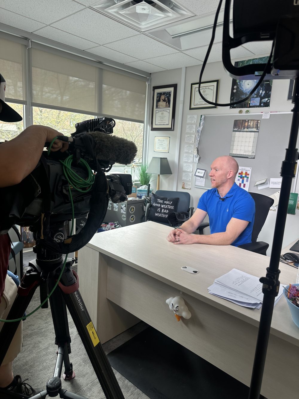 Vertical image of Terry Shirley facing a camera while sitting at his desk in his office. Weather memorabilia can be seen on his walls and desk.