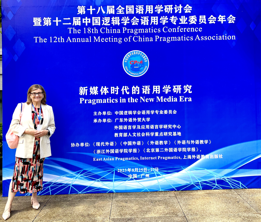 Blitvich standing in from of a large blue billboard that says "The 18th China Pragmatics Conference" and "The 12th Annual Meeting of the China Pragmatics Association"