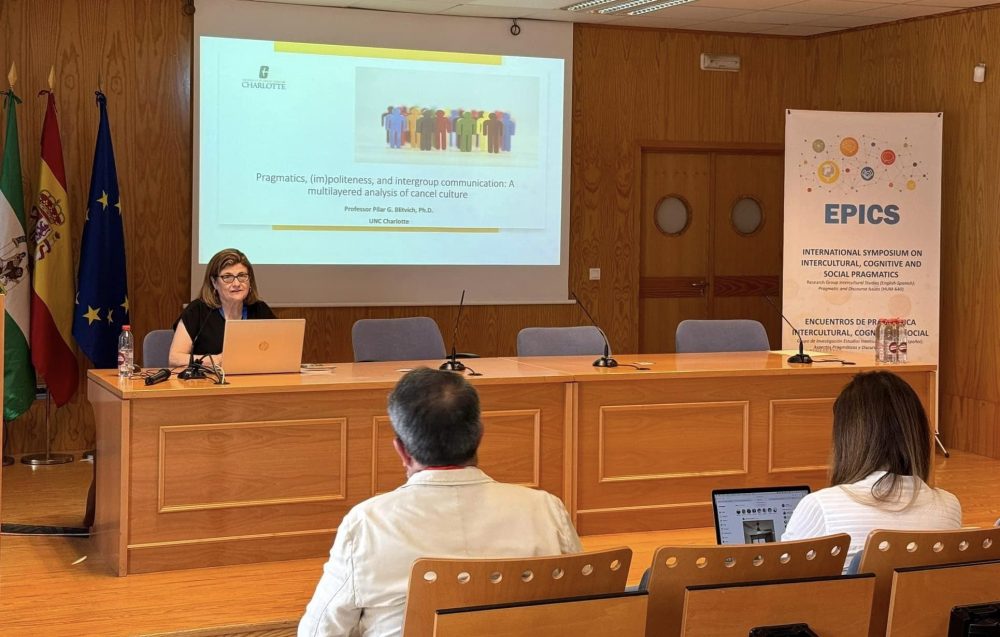 Blitvich sits on the left behind a large table with a presentation behind her. Two audience members are in the foreground, and an "EPICS" banner is on the right side.