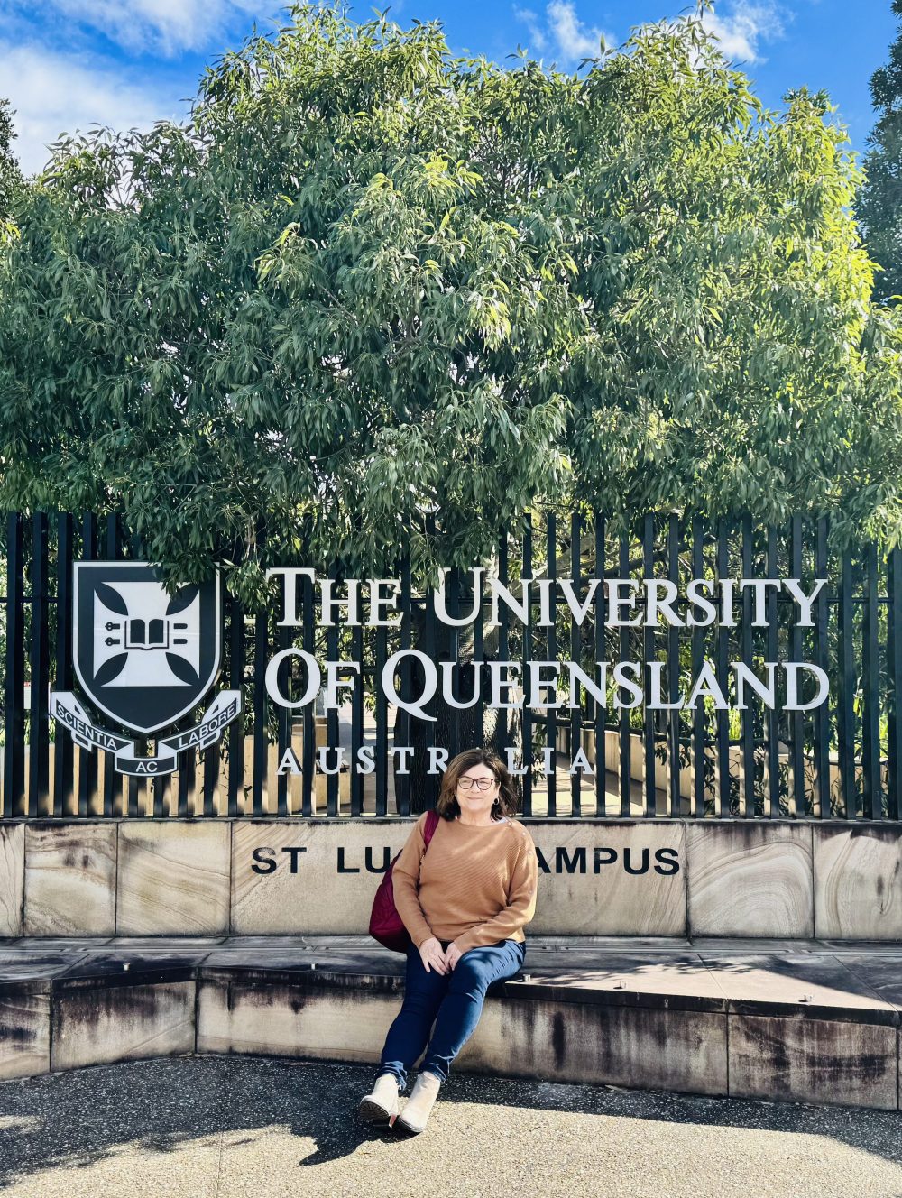 Blitvich sitting on a step in front of a large logo and a sign that says "The University of Queensland Australia"
