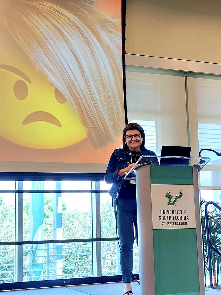 Blitvich standing behind a podium that says "University of South Florida St. Petersburg" on stage in front of a presentation with an angry emoji.