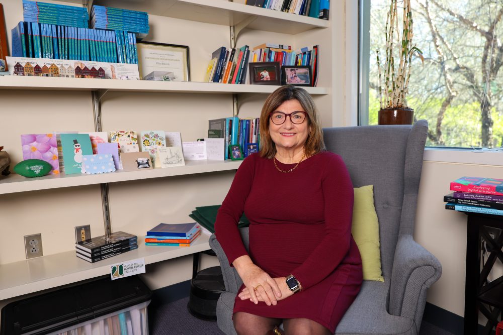 Blitvich sitting on a chair in the corner of her office in front of a window and her bookshelves. Copies of her books and journals are next to her.