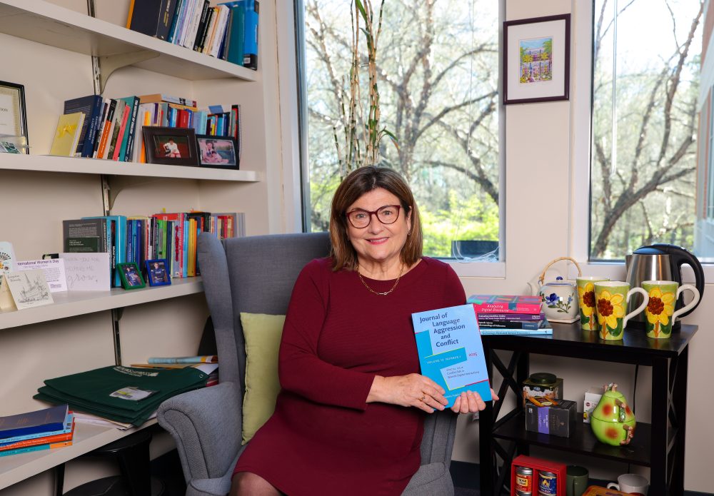 Blitvich sitting on a chair in the corner of her office, holding a copy of the Journal of Language Aggression and Conflict