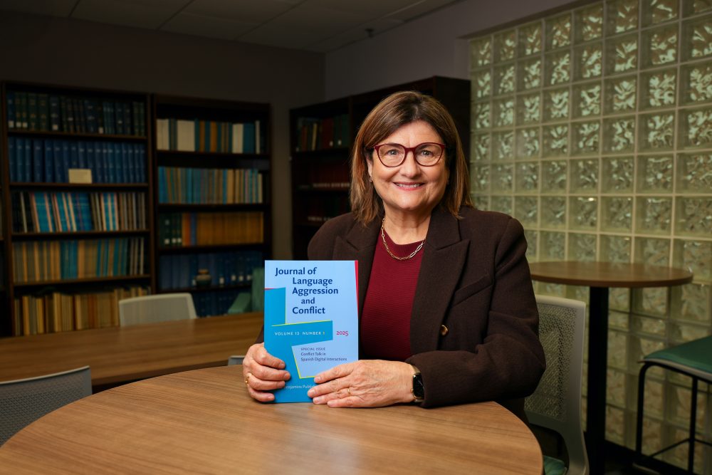 Blitvich sitting at a table with bookshelves behind her. She is holding a 2025 volume of the "Journal of Language Aggression and Conflict"
