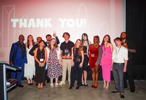 A group of professionally dressed students and their advisor at the Independent Picture House at the end of the Gold Reel Film Festival. The movie screen behind them says "Thank You!"