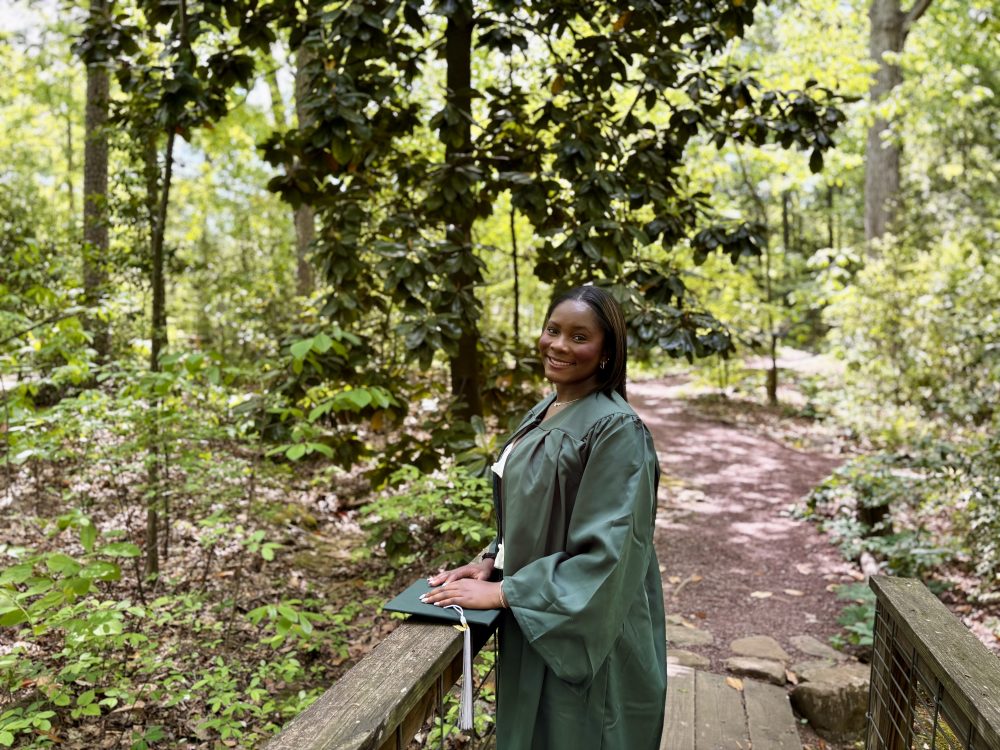 Frink poses in graduation gown on a wooden bridge, with a path and trees in the background.