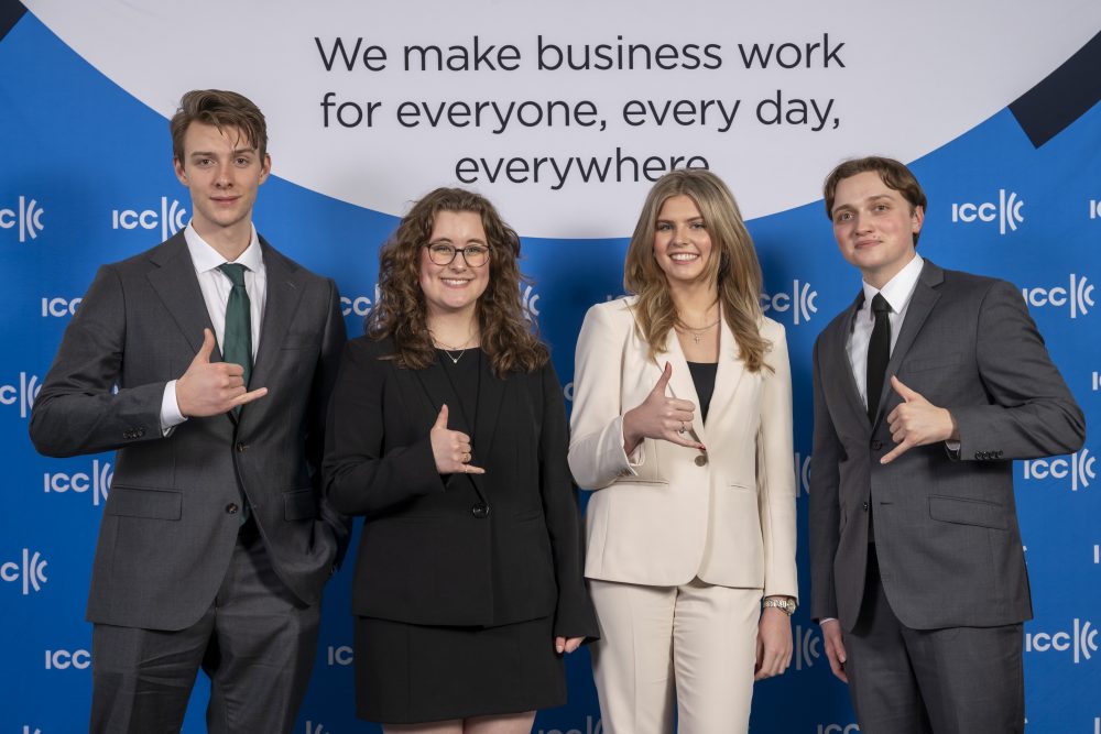 The four students pose in front of the ICC banner, smiling and doing the UNC Charlotte pickaxe with their hands.
