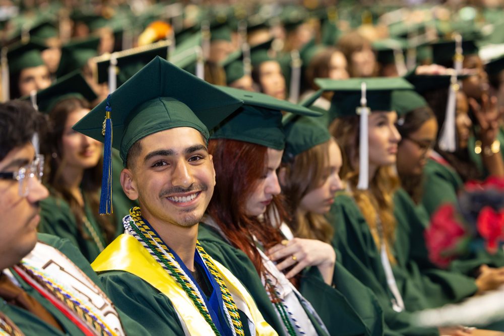 Jude Bianouni seated in the crowd of UNC Charlotte graduates smiles at the camera.