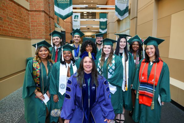 A group of smiling UNC Charlotte graduates fron the College of Humanities & Earth and Social Sciences in regalia with faculty marshal Jeanette Bennett.