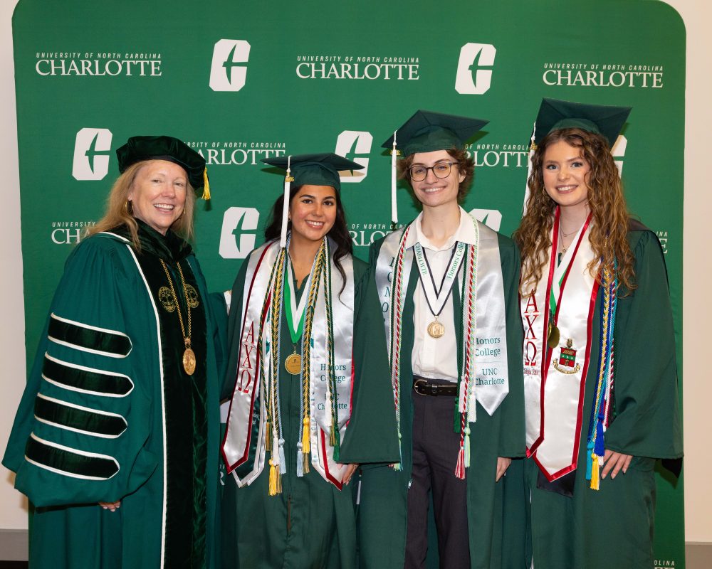 Four people wearing UNC Charlotte graduation regalia in front of a backdrop. UNC Charlotte Chancellor Sharon L. Gaber, Senior Class President Emily Hoyle, student commencement speaker Foster Duckworth, bell ringer Kierra Andrews.