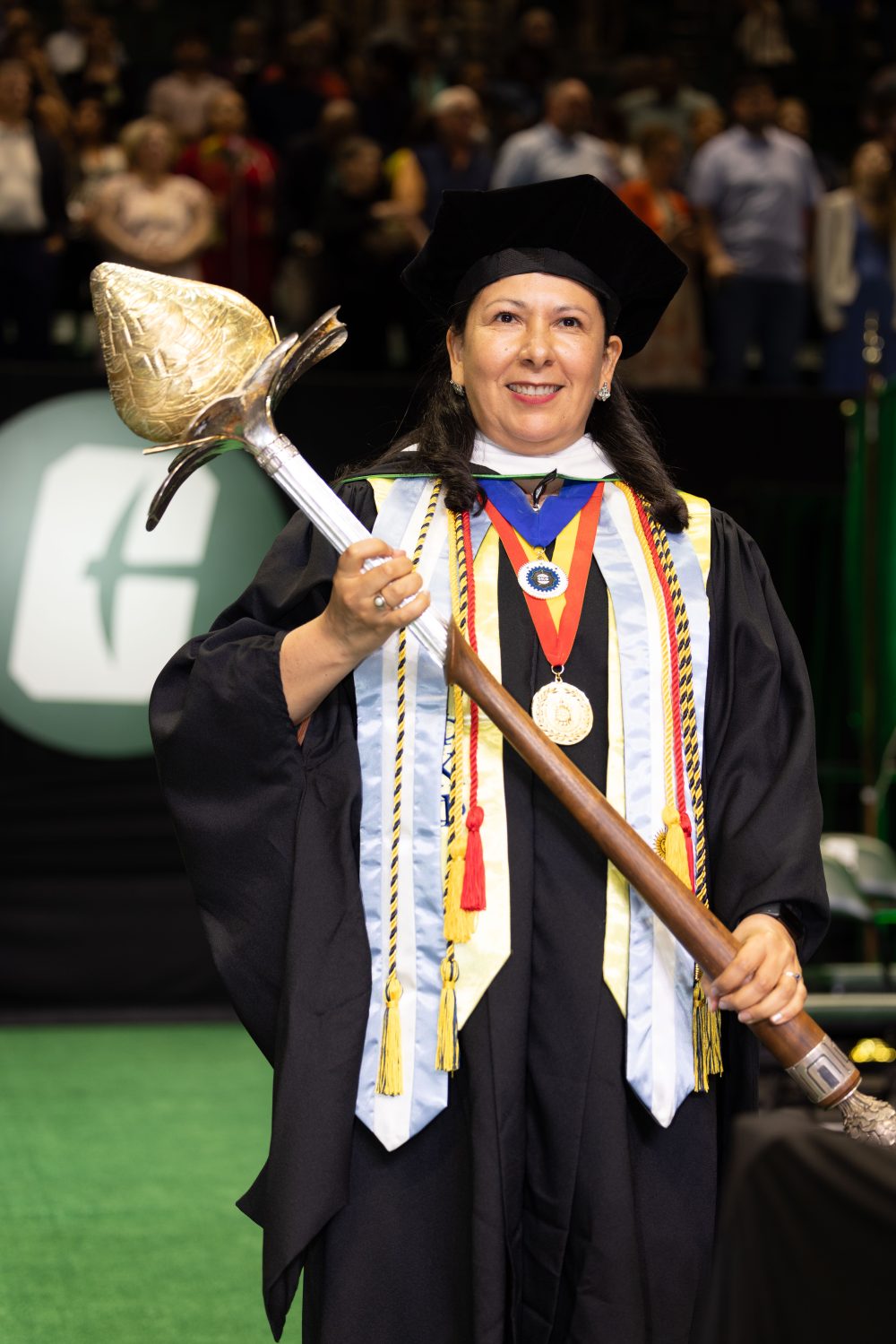 UNC Charlotte's Susana Cisneros wears graduation regalia on stage at the commencement ceremony, and holds the ceremonial mace. At the base is a gold pine cone. Moving upward into the native walnut handle is one of Charlotte’s former logos. The upper silver section is carved to represent the Belk Tower that once stood in Belk Plaza, which then blossoms into a dogwood flower with a gold-plated hornet’s nest.
