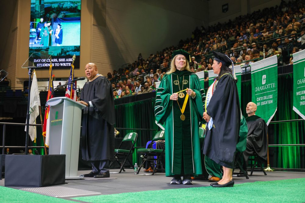 UNC Board of Governor's member Joel Ford speaks at the UNC Charlotte podium on stage at graduation while Chancellor Sharon L. Gaber holds the medal for Excellence in Teaching, waiting to put it on Susana Cisneros.
