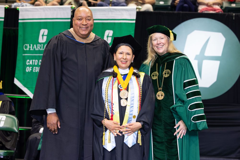 Three people dressed in graduation regalia on stage at UNC Charlotte's graduation ceremony.