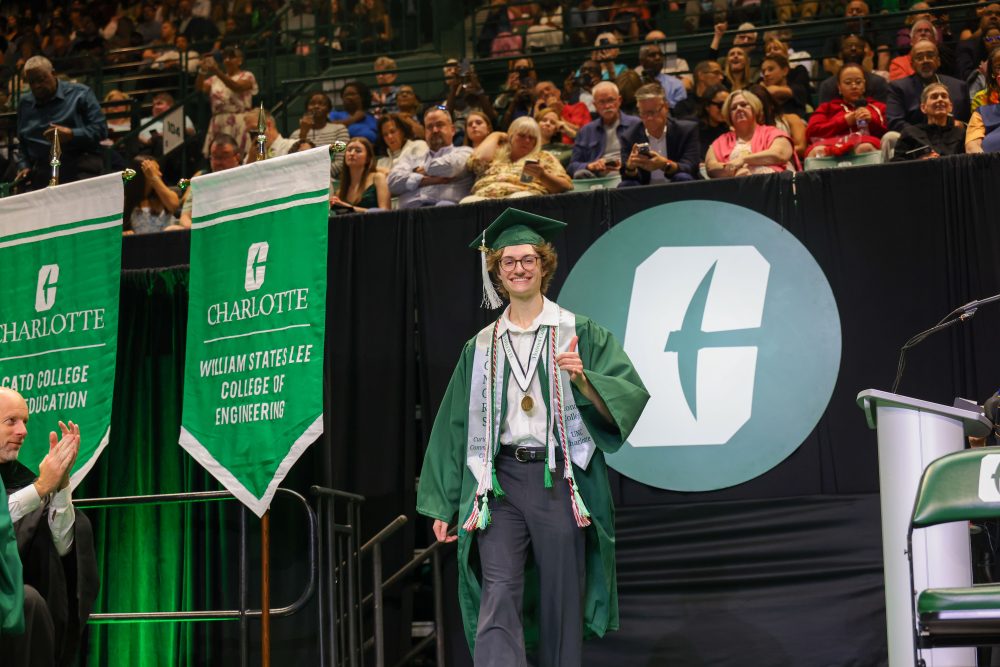 Foster Duckworth crosses the stage in graduation regalia at UNC Charlotte's commencement and smiles, showing the pickaxe hand sign.
