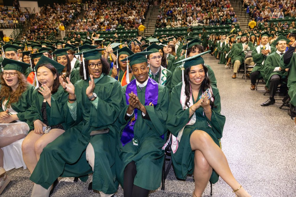 A group of Niner graduates in the audience wearing UNC Charlotte graduation regalia and clapping and smiling.