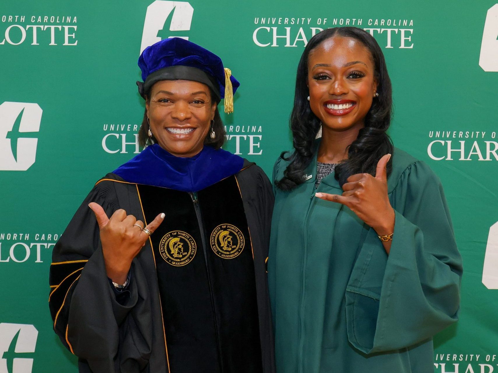 Two people wearing UNC Charlotte graduation regalia in front of a backdrop, holding up Niner pickaxe hand signs. Faculty Senate President Debra Smith, SGA president Madison Brown.