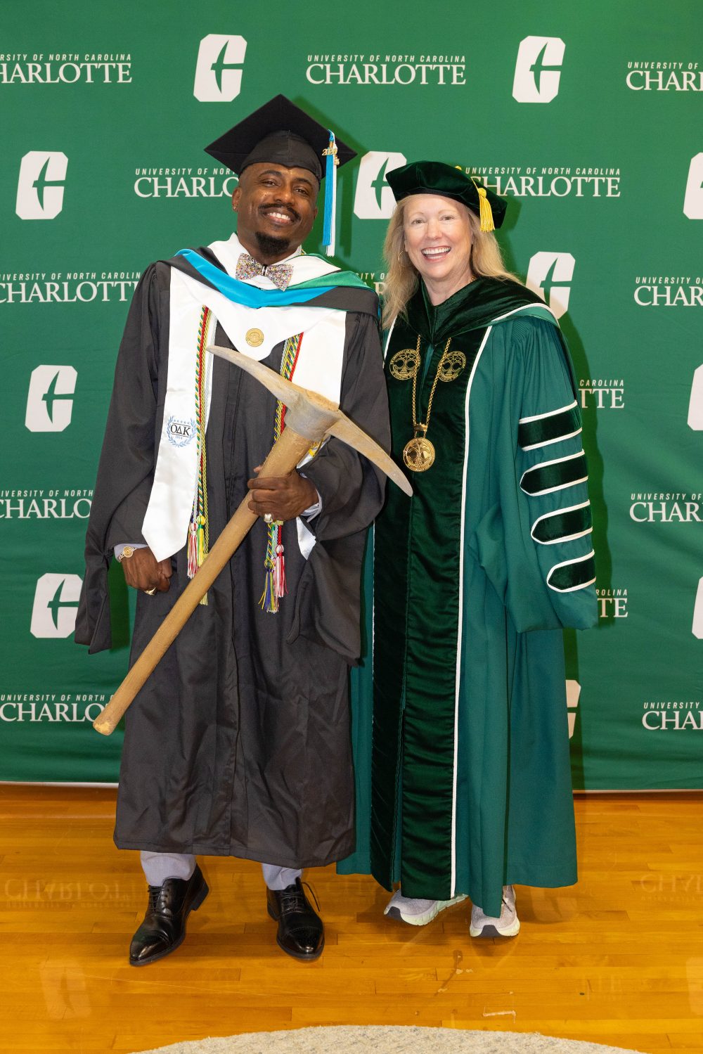 S.L. Ivey wears his graduation regalia and holds the gold pickaxe he was awarded as the Golden Niner, standing with UNC Charlotte Chancellor Sharon L. Gaber.