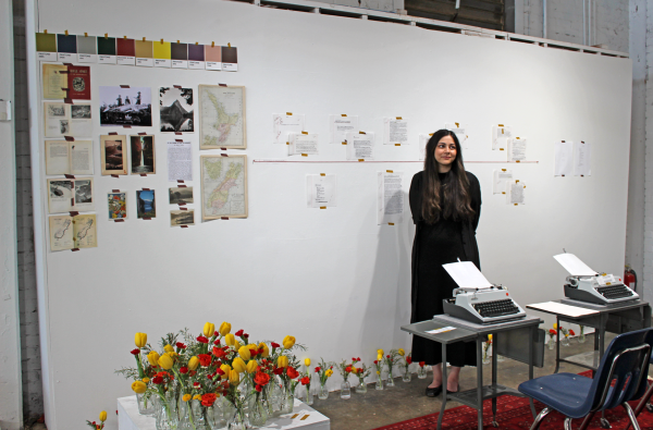 E.E. Hussey stands in her showcase space in front of a white wall covered with early drafts, archival materials and first‑draft typewriter pages. In the foreground are two of her typewriters and vases of flowers.