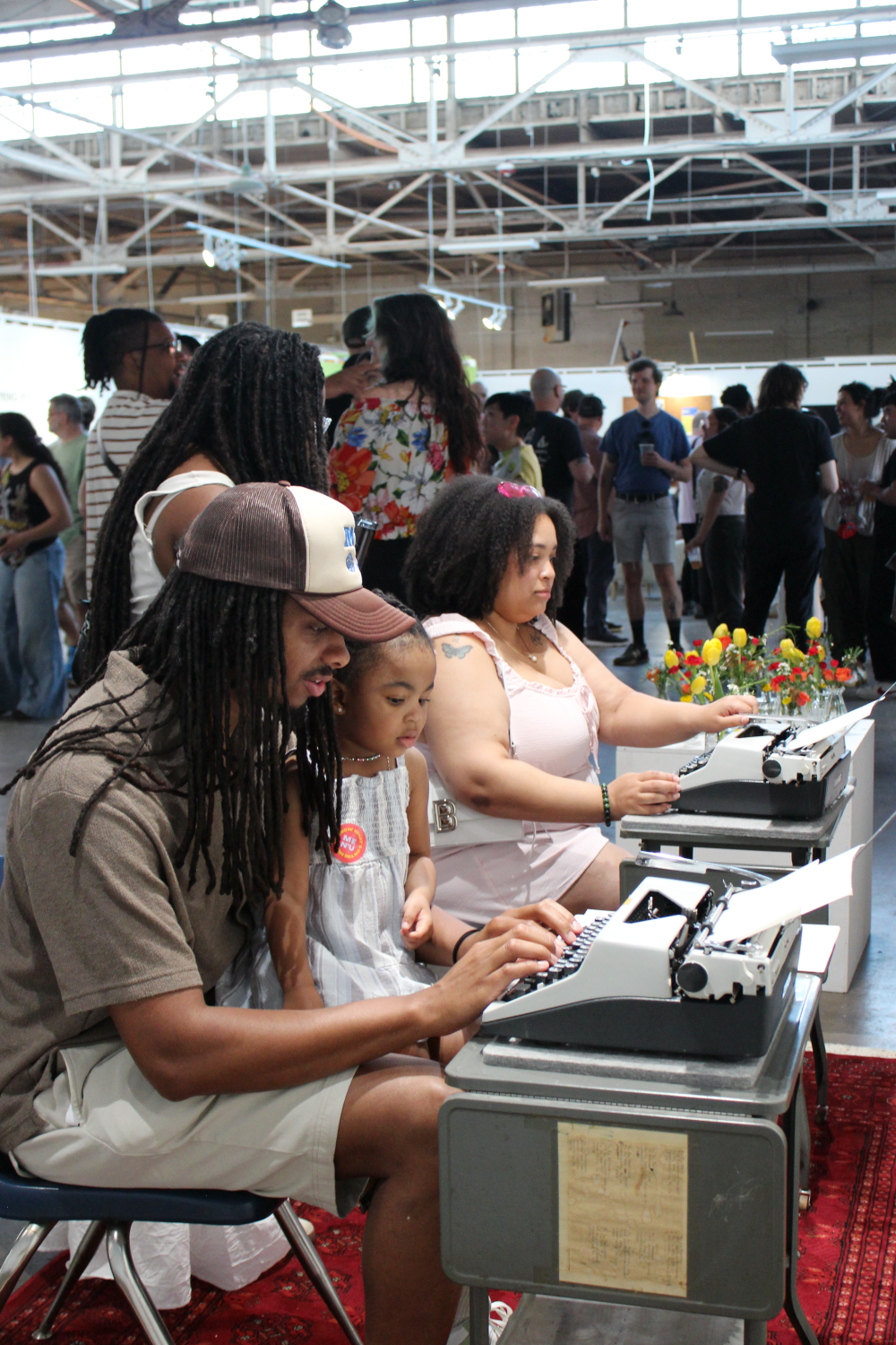 Attendees sitting at the typewriters. A dad and his daughter sit together, with two women in the background. Attendees stand and talk around them. 