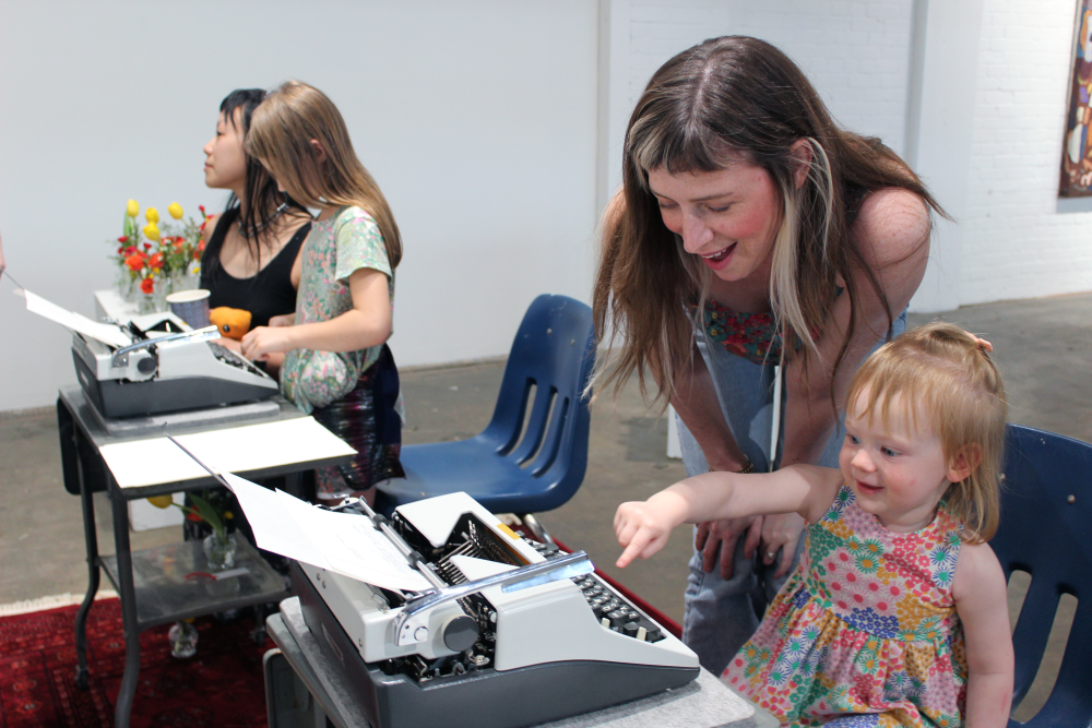 A woman smiles down at a child using one finger to type on the typewriter.