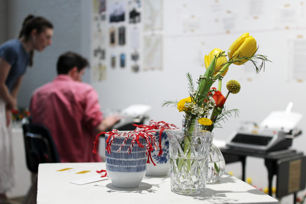 Close up of flower vases and bookmarks sitting on a pedestal. The gallery can be seen in the background but blurred, with a man typing on a typewriter.