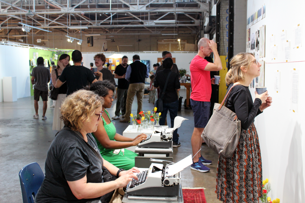 Chancellor types on the typewriter while Hutchcraft looks at the gallery wall.