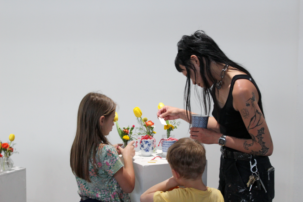Vu holding a bookmark that was on a podium containing flower vases and two cups filled with bookmarks. Two children look on.