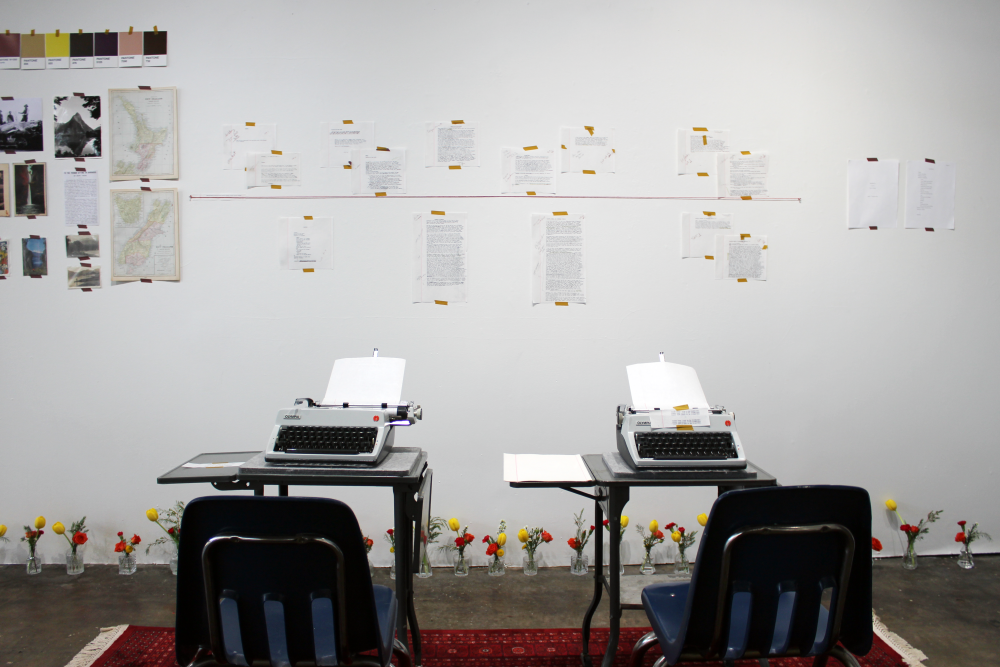 The gallery empty before opening. Two typewriters on vintage desks, with flower vases on the floor and a gallery wall filled with photos, maps, color swatches and early drafts.