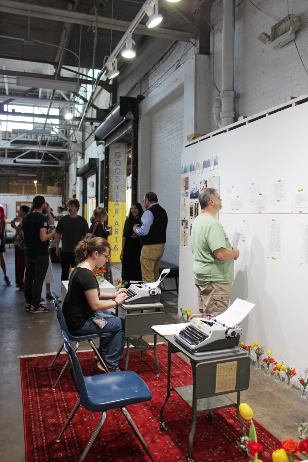 A man looks at the writings on the wall as a woman types on the typewriter. Hussey and colleagues speak in the background where you can see the "Goodyear Arts" painted on the entrance of the space.