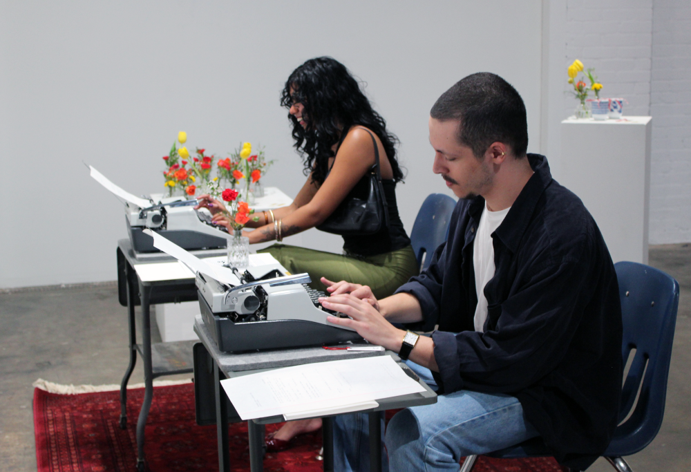 Beets types on a vintage typewriter. A woman smiles as she types on the other typewriter next to his, with flower vases in the background.