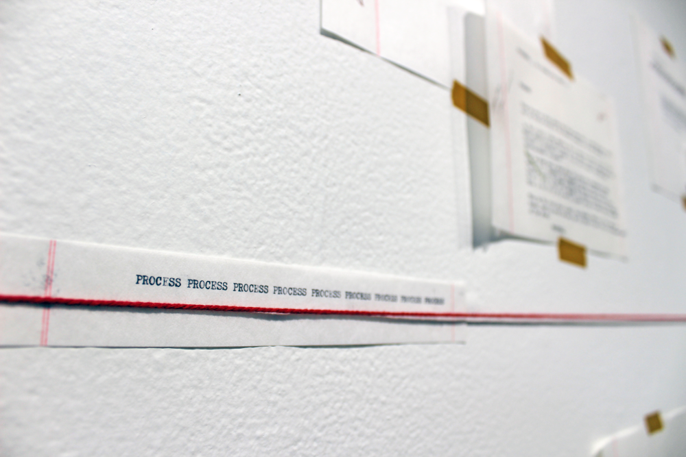 A close up of a sliver of text on the gallery wall which reads, "PROCESS" 9 times. A red string spans across showing a timeline of work.